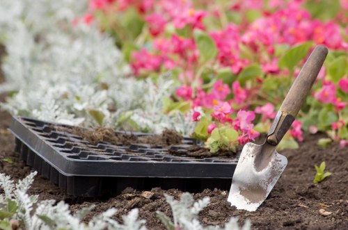 A gardening scene showing a rectangular black seed tray placed on freshly turned dark soil in a garden, with a small hand trowel with a wooden handle partially inserted into the ground. Surrounding the tray are vibrant pink and white flowering plants, creating a colourful backdrop. The soil appears moist and well-prepared for planting, and the background features a mix of green foliage and flowering shrubs, suggesting a well-maintained outdoor space typical of gardens in Southwark. The natural lighting indicates a bright day, ideal for outdoor gardening activities. This image illustrates typical elements of garden planting and bed preparation, aligning with residential gardening and landscaping services offered by Lawn Mowing Southwark.