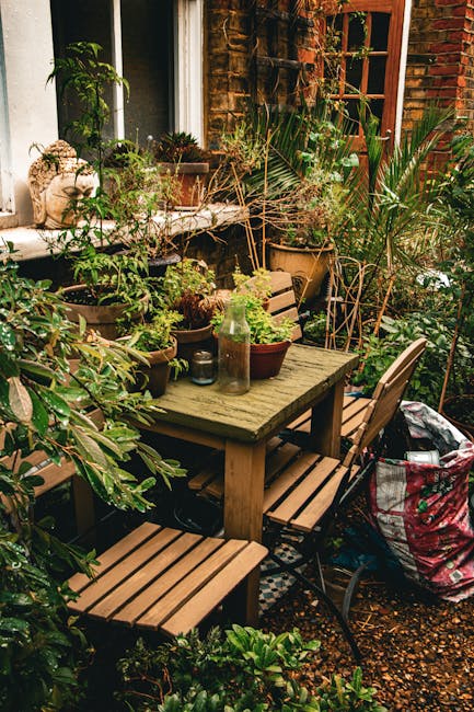 A small, urban garden space featuring a weathered wooden table surrounded by four matching wooden and metal chairs, set against a brick wall with a window. The table is covered with an assortment of p